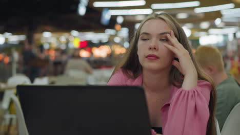 young woman in pink dress sits in front of her laptop, placing her hand on her head while blinking and appearing deep in thought, blurred background of a busy, well-lit restaurant with blur light