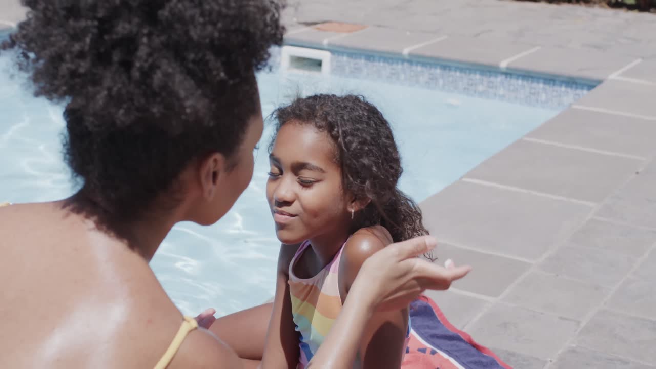 Happy african american mother and daughter applying sunscreen by swimming pool, slow motion