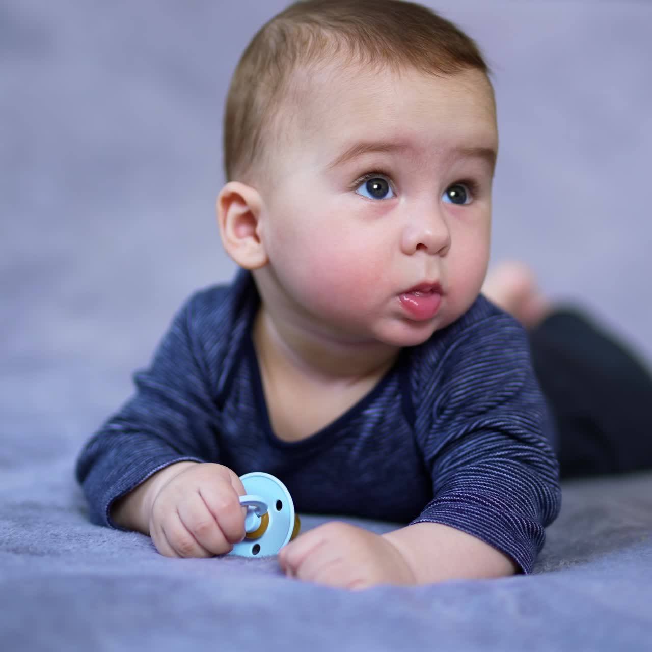 Sweet tiny boy lies on bed holding a pacifier in his hand. Beautiful kid turns his head to look around and smiles adorably. Close up