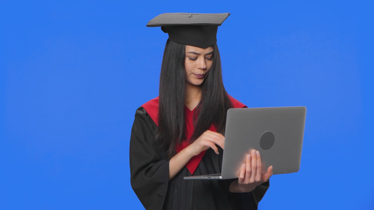 Ritratto di studentessa in toga e tocco da laureata, che comunica online usando un laptop. Giovane donna in posa in studio con sfondo a schermo blu. Primo piano. Pronto per slow motion 59.94fps