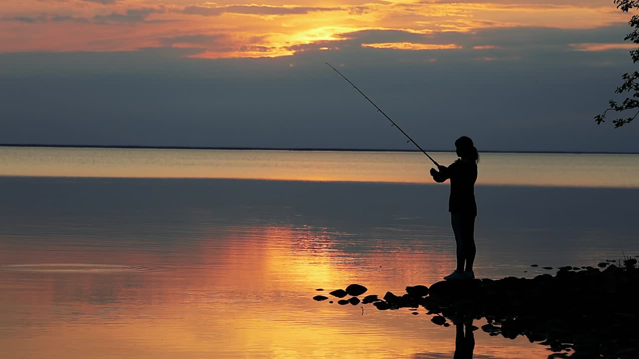 Wanita memancing dengan joran pancing berputar dengan latar belakang matahari terbenam.