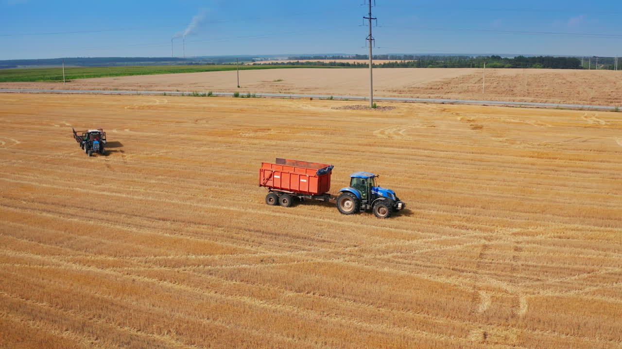 Tractor machines moving by the agricultural plantation. Truck being uploaded with the hay bales at the background.