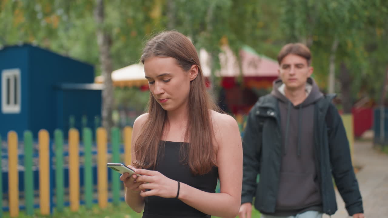 Bílý teenager píše zprávu v zábavním parku, student čeká na telefonu, rozptýlen zprávami, v pozadí plot parku a kolotoč, jemný nervózní výraz, ležérní oblečení a batoh naznačují školní život