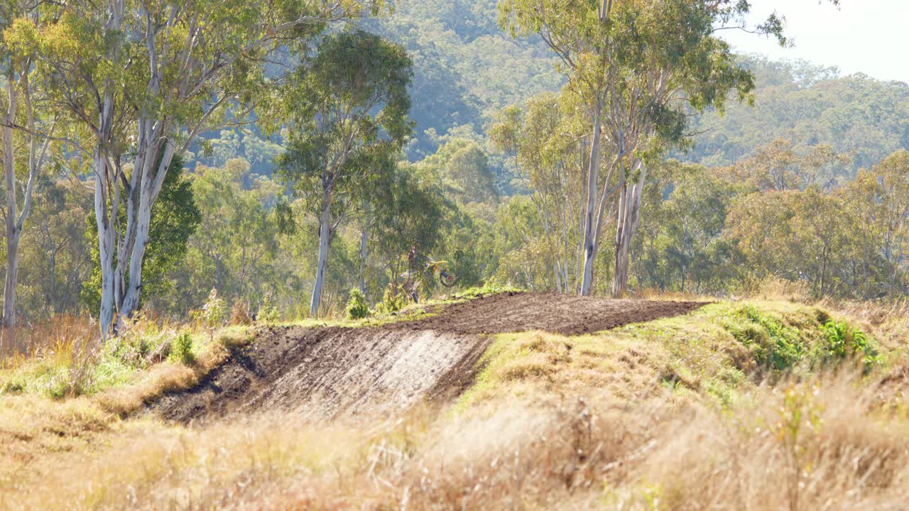 A dirt bike rider approaches a dirt jump on an outdoor motocross track surrounded by dry grass and eucalyptus trees under natural daylight with a distant, static camera view