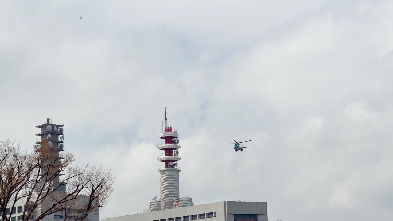 Et helikopter flyr over en overskyet himmel forbi moderne telekommunikasjonstårn og urban arkitektur.