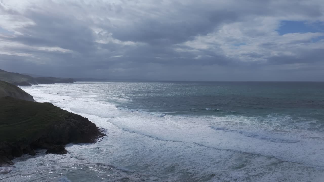 High-altitude drone orbit in slow motion shows the turquoise Cantabrian Sea meeting reddish-brown cliffs under a cloudy sky with light rays breaking through, forming mist and soft marine textures