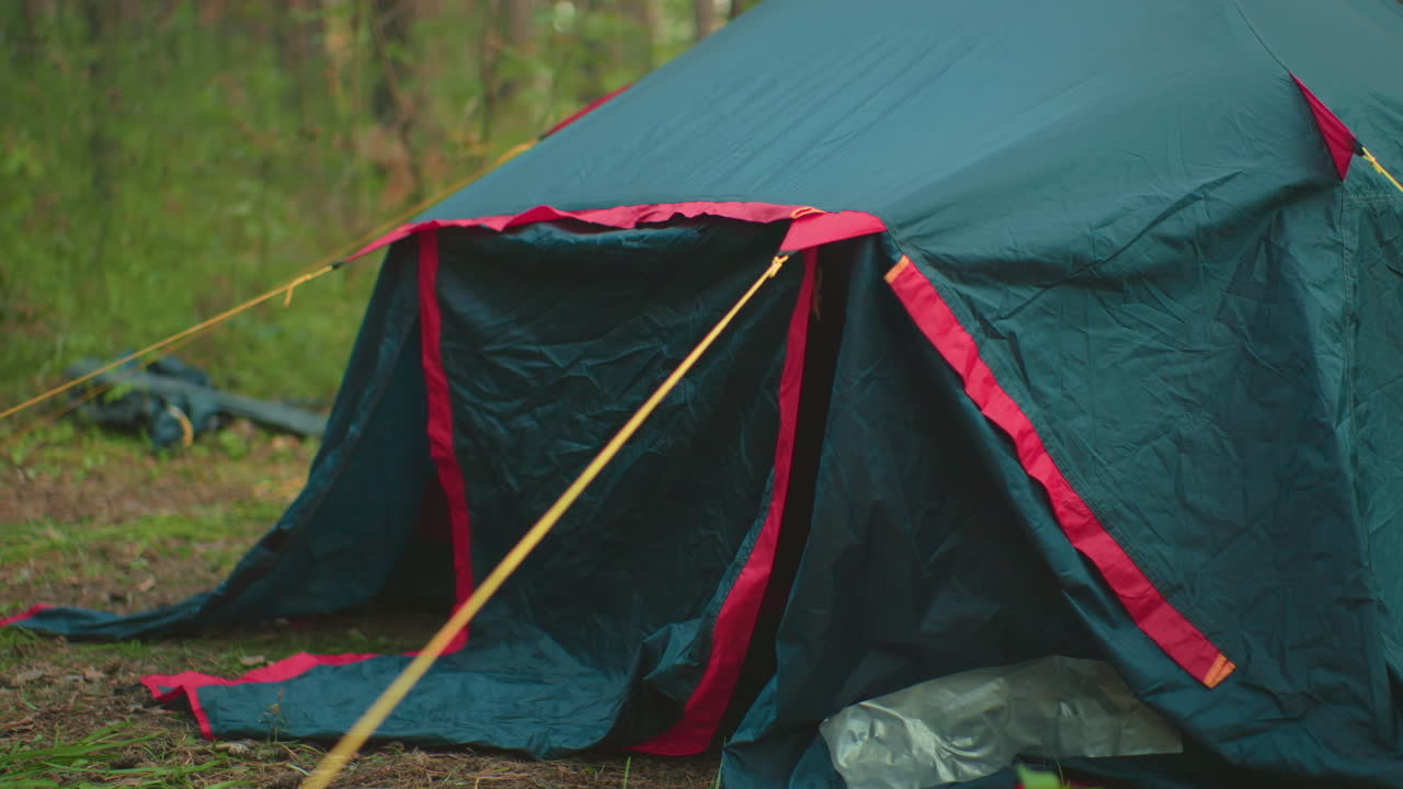 Close up of forest camping tent partially open, secured with bright yellow ropes and red trim, resting on ground among grass and soil, illustrating outdoor shelter setup in natural environment