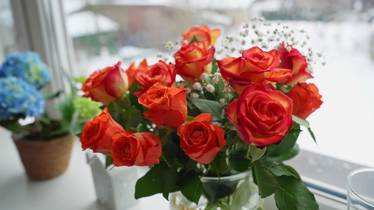 明るい窓辺に置かれたガラスの花瓶に入ったオレンジ色のバラの花束。窓の外には柔らかな冬のボケが広がる。ロマンスやお祝いのメッセージに使えるコピースペースのある、居心地の良いホームデコのフローラルシーン。