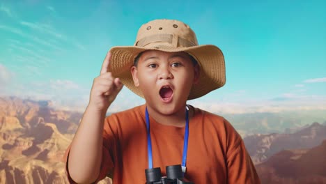 asian boy with a hat and binoculars thinking and looking around then raising his index finger while traveling at the top of mountain. boy researcher, travel adventure concept, close up