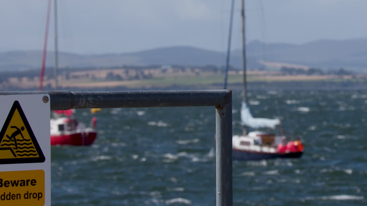 Windy coastal pier scene with warning sign, choppy sea, moving sailboats, and distant hills