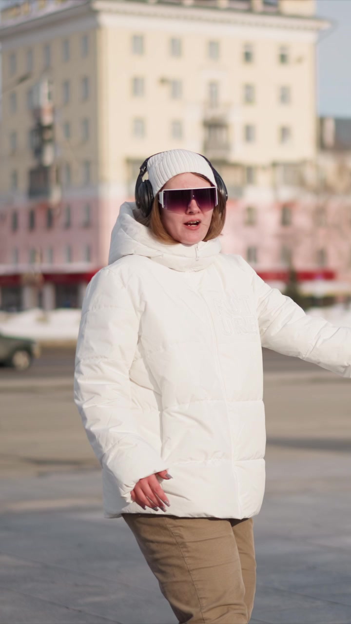 Woman wearing white puffer jacket black headphones and brown pants walks outdoors in urban setting with buildings visible. Woman gestures naturally as woman moves through environment