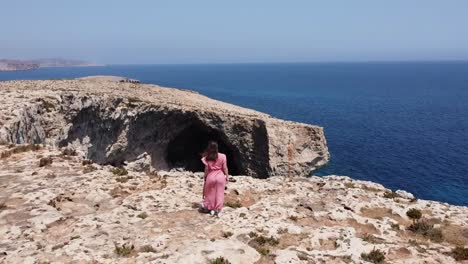 Aerial-drone-view-of-a-woman-with-a-long-summer-dress-walking-near-the-cliff,-with-the-wind,-traveling-in-Malta,-Europe
