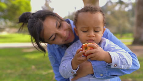 Mixed-race-pretty-mother-hugging-son,-looking-him-eating-apple