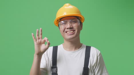 close up of asian man worker wearing goggles and safety helmet smiling and showing okay gesture to camera while standing in the green screen background studio