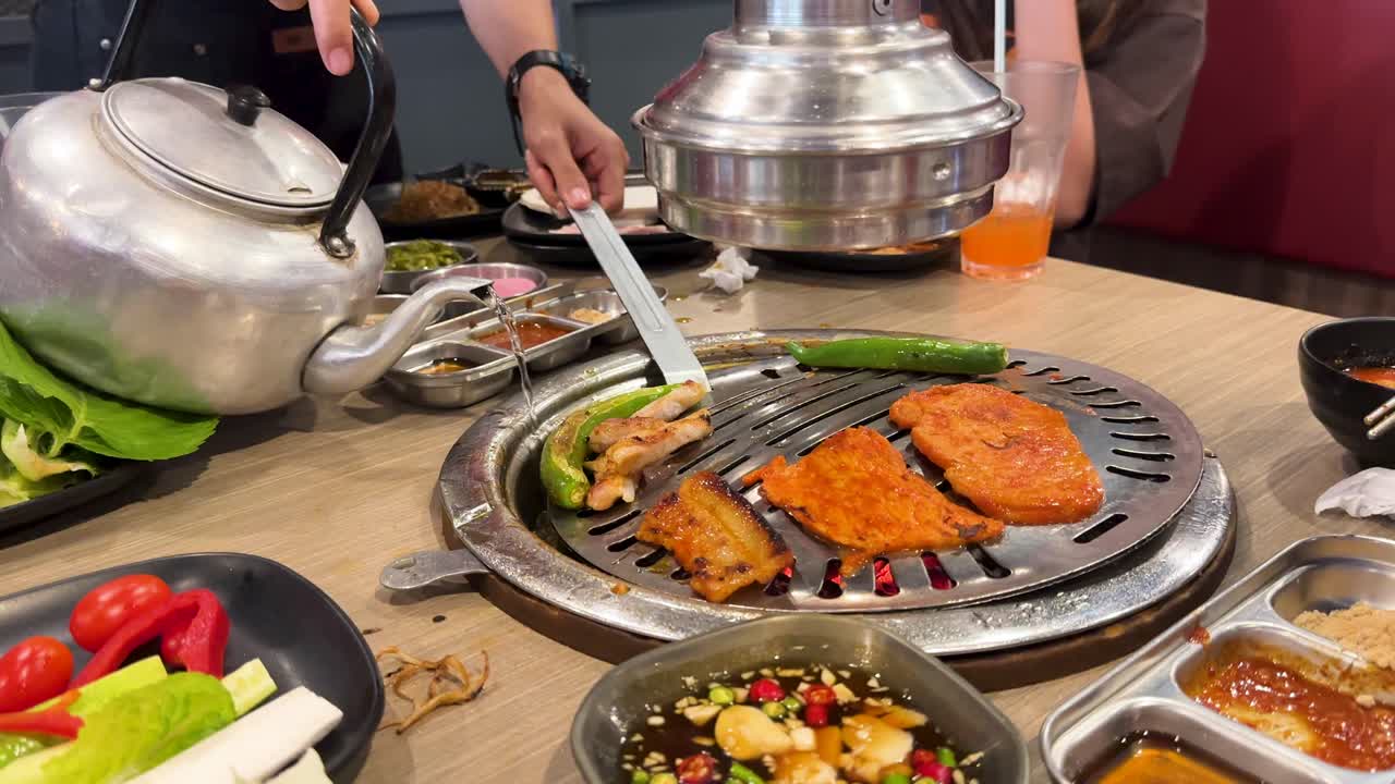 Grilled pork and vegetables on a Korean BBQ grill in a Bangkok restaurant. Bright lighting, vibrant colors, and interactive dining atmosphere