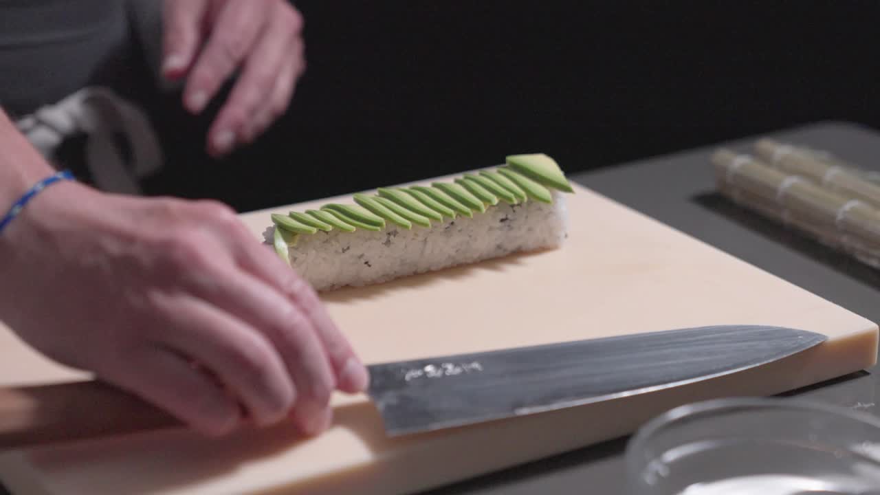 A roaming shot captures a sushi chef placing fresh avocado slices onto a sushi roll, showcasing the precision and artistry of sushi preparation. Ideal for culinary and sushi-making content.