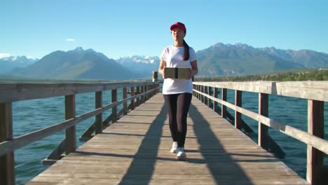 a woman strolls along a wooden pier, carrying a package while enjoying a clear sunny day. the surrounding mountains provide a stunning backdrop to her outdoor experience.