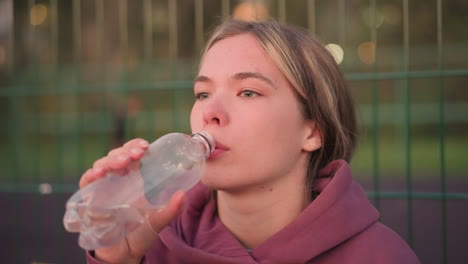 young lady in maroon hoodie drinking water from a bottle after workout, captured in side profile, with blurred bar fence in background, refreshing moment post-exercise