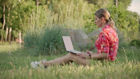 a middle-aged woman sits on the grass in the park. using a video link on a laptop, she conducts a conversation.