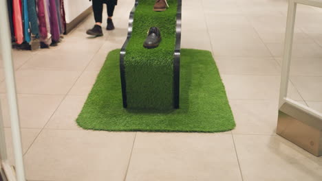 a black and brown shoe placed on a unique stand covered in green artificial grass. the display is part of a retail store setup with clothing visible in the background and a person walking past