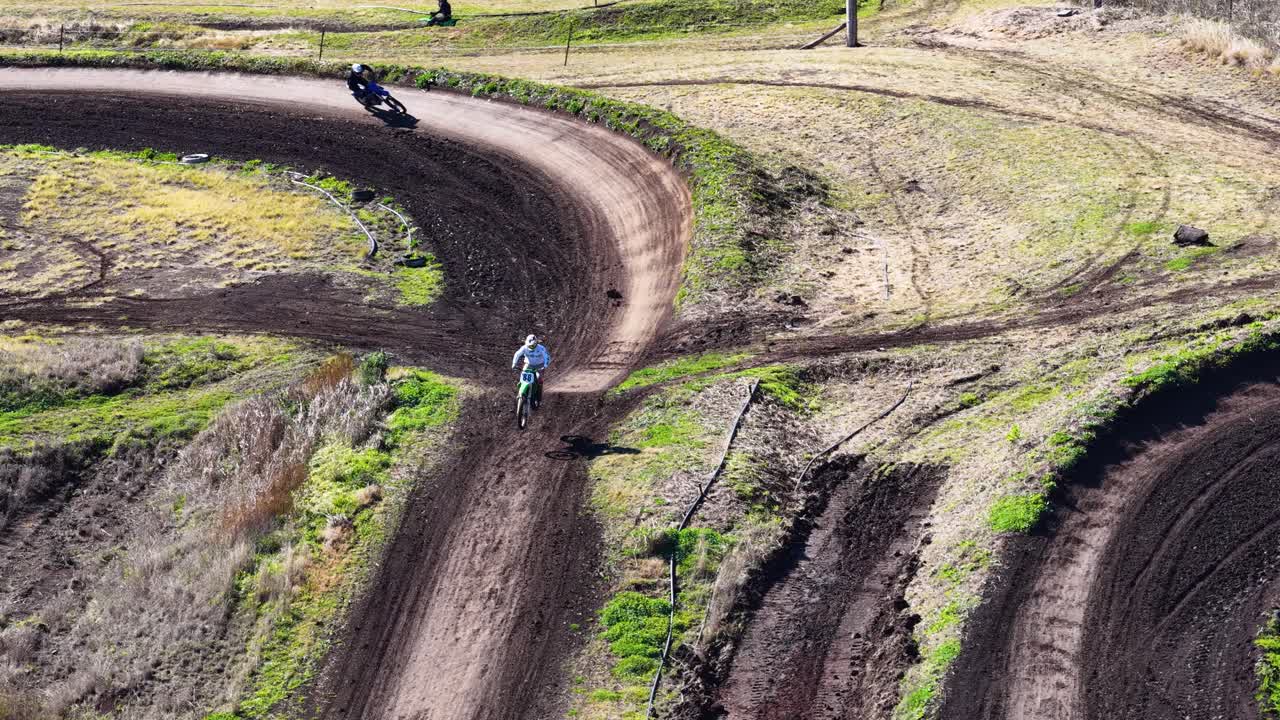 A motocross rider in full gear rides a dirt bike along a winding, sunlit track surrounded by bushland, captured from a high aerial perspective