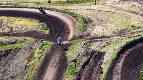 a motocross rider in full gear rides a dirt bike along a winding, sunlit track surrounded by bushland, captured from a high aerial perspective
