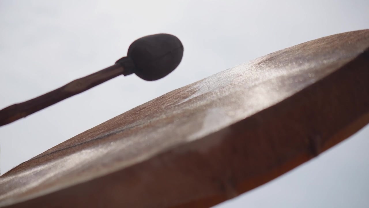 Low-angle close-up of a musician striking a traditional frame drum with a padded beater, capturing rhythmic movement during an acoustic percussion performance.