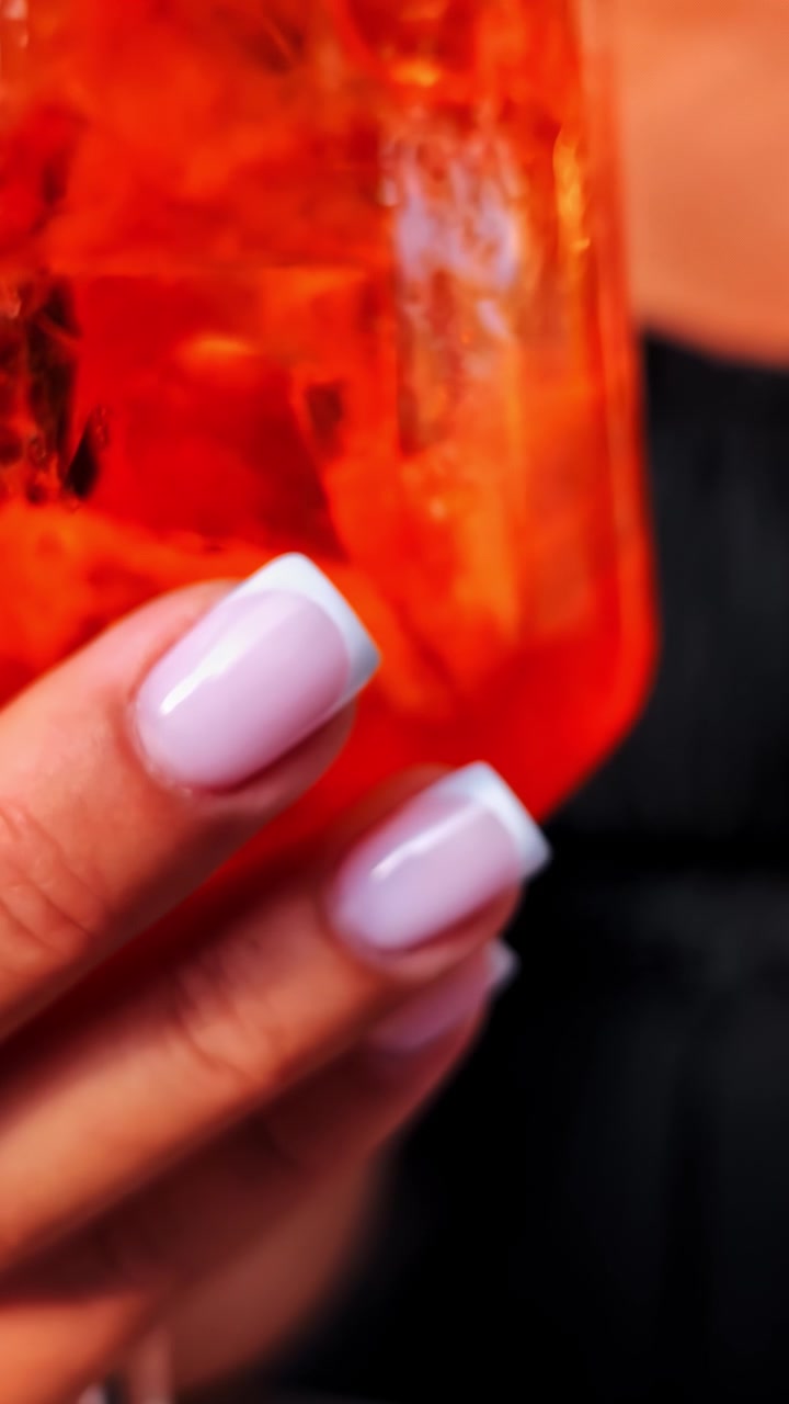 Hånd der holder en afkølet orange cocktail med isterninger, vertikalt detaljeret nærbillede.