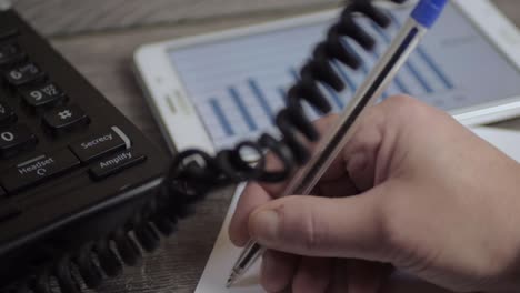 Busy person in phone conversation writing notes while looking at graph on a tablet