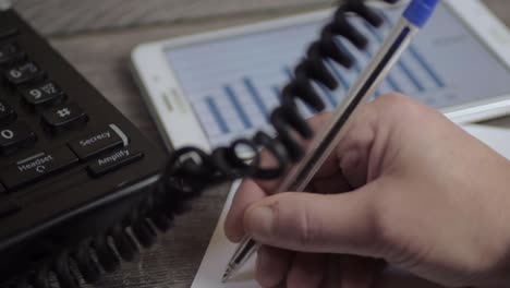 busy person in phone conversation writing notes while looking at graph on a tablet