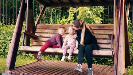 young pretty mother with children swinging on a wooden swing near the summer house