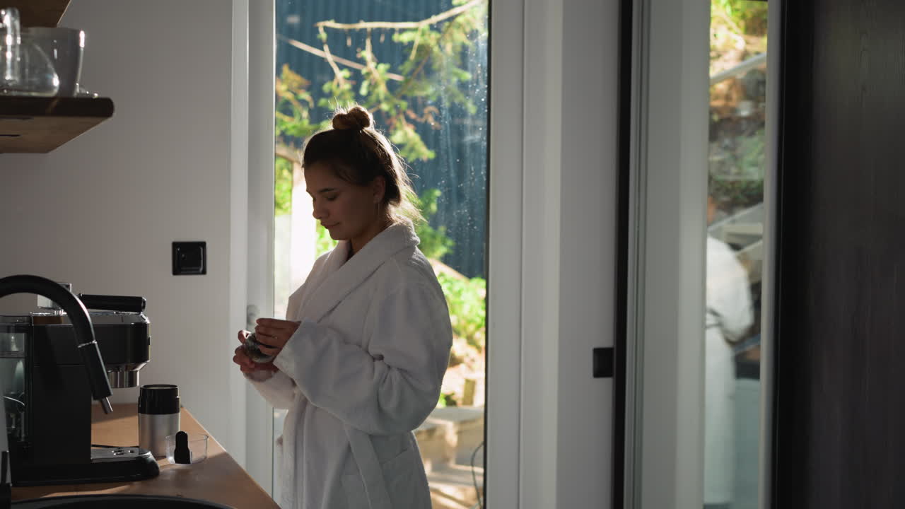 Woman enjoys aroma of coffee beans in kitchen. Lady in terry bathrobe smells coffee grains from jar enjoying early morning. Relaxing morning moment