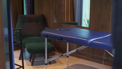 mesa de masaje vacía en una habitación de spa de un hotel, acompañada de un sillón y una mesa pequeña. un espejo refleja plantas exuberantes, mejorando el ambiente sereno y relajante