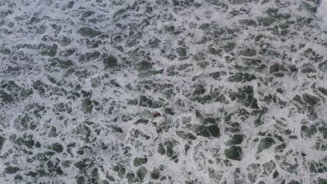 Surfers-paddling-through-breaking-waves-in-Pacific-Ocean,-aerial-bird's-eye-view