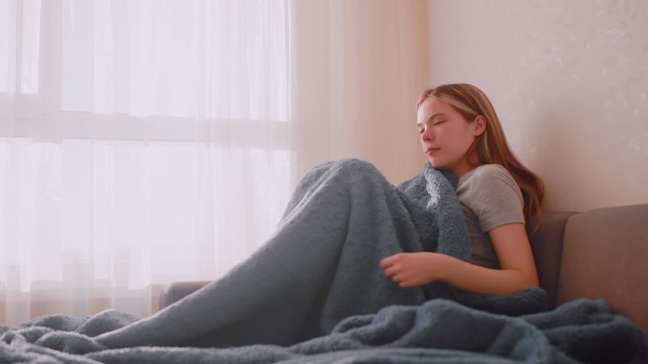 Young woman seated on sofa covering herself with soft blanket near bright window as sunlight passes through curtain creating indoor mood of comfort warmth