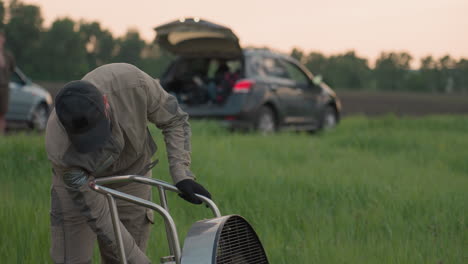 man in cap and gloves preparing large fan beside hot air balloon basket on grassy field at sunset wind rustling grass in gentle evening light holding sturdy metal frame in serene rural landscape