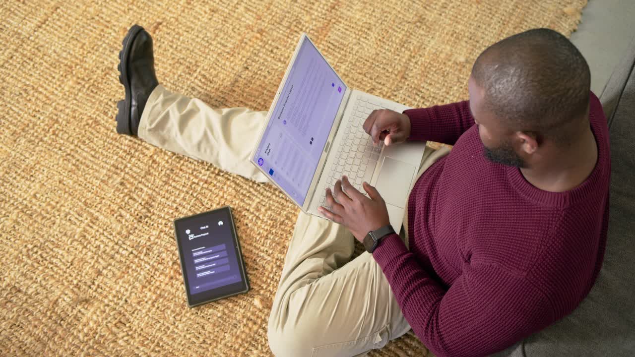As clip begins man typing on laptop atop rug and glancing at tablet to complete work