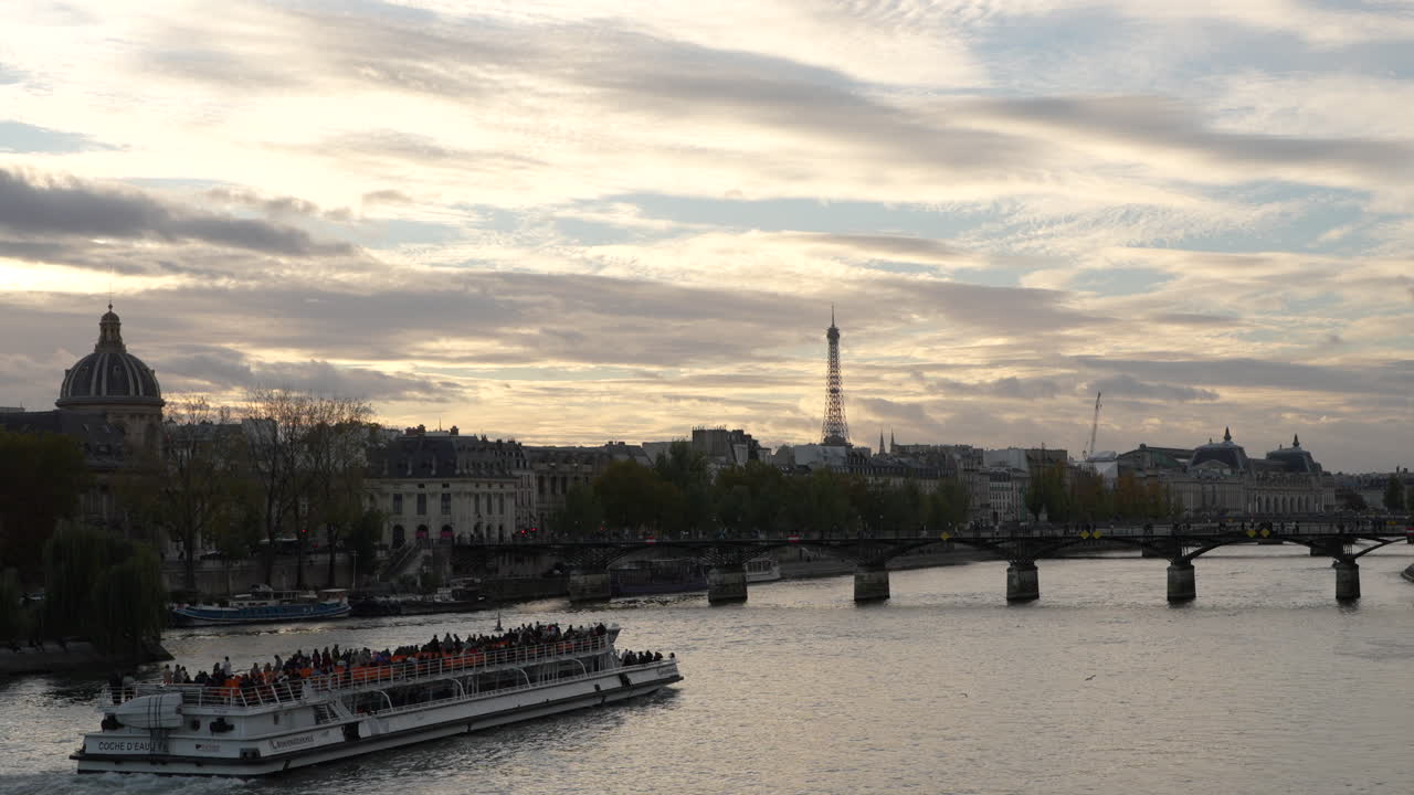 Wide shot of the Pont des Arts bridge and Eiffel Tower at sunset in Paris. A tourist cruise boat sails along the Seine River under dramatic clouds, the city’s romantic skyline and evening atmosphere