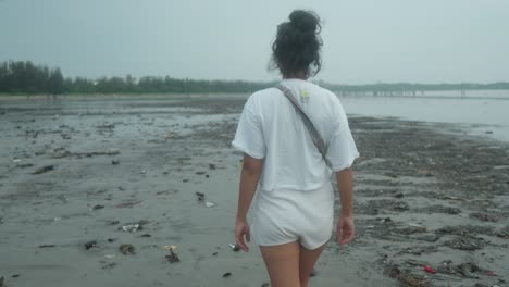 medium dolly shot of a young indian woman in white shirt and fanny pack while taking an evening walk on the beach during her summer vacation in slow motion