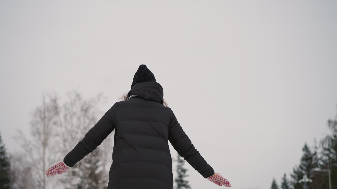 Athletic girl in black winter coat and knit hat with colorful mittens seen from behind outdoors in snowy environment, arms extended for balance, expressing freedom, energy in wintertime
