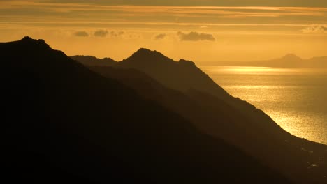 aerial cinematic shot of majestic mountain silhouettes backed by brilliant refection of the sun on the ocean and a distant mountain