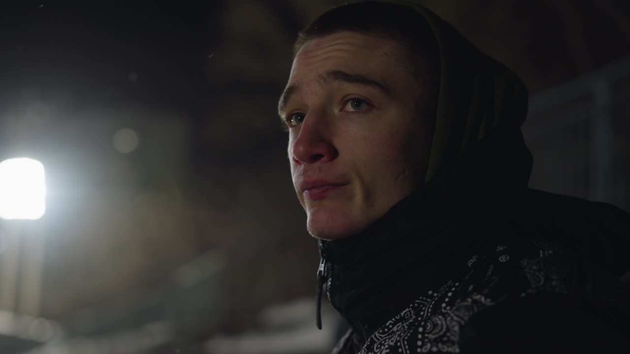 Side profile of young man wearing green hoodie and winter jacket looking contemplatively into distance during nighttime, under dim ambient lighting with cold atmosphere and muted background setting