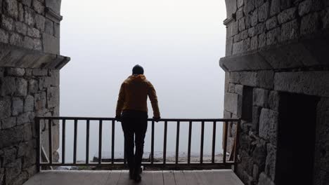 Woman-walks-to-railing-in-old-church,-looking-out-to-foggy-day-outside