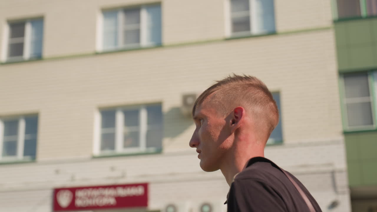 Young Caucasian Man Walking Past Apartment Building, Side Profile, Shaved Haircut, Earpiece Visible, Sunlit Facade, Tense Posture, Pacing Stride, Subtle Paranoia And Ocd Tension In Expression