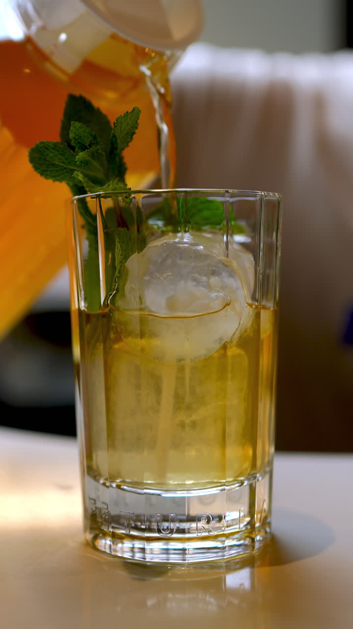 Vertical footage A bartender pours iced tea from a jug into a glass filled with ice and decorated with a sprig of mint, creating a refreshing summer beverage