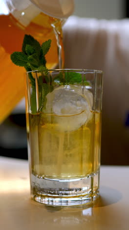 vertical footage a bartender pours iced tea from a jug into a glass filled with ice and decorated with a sprig of mint, creating a refreshing summer beverage
