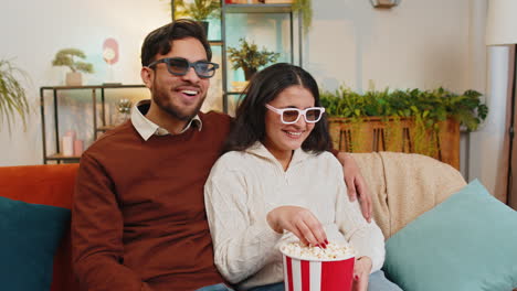 cheerful young indian couple in 3d glasses eating popcorn and watching comedy movie sitting on sofa
