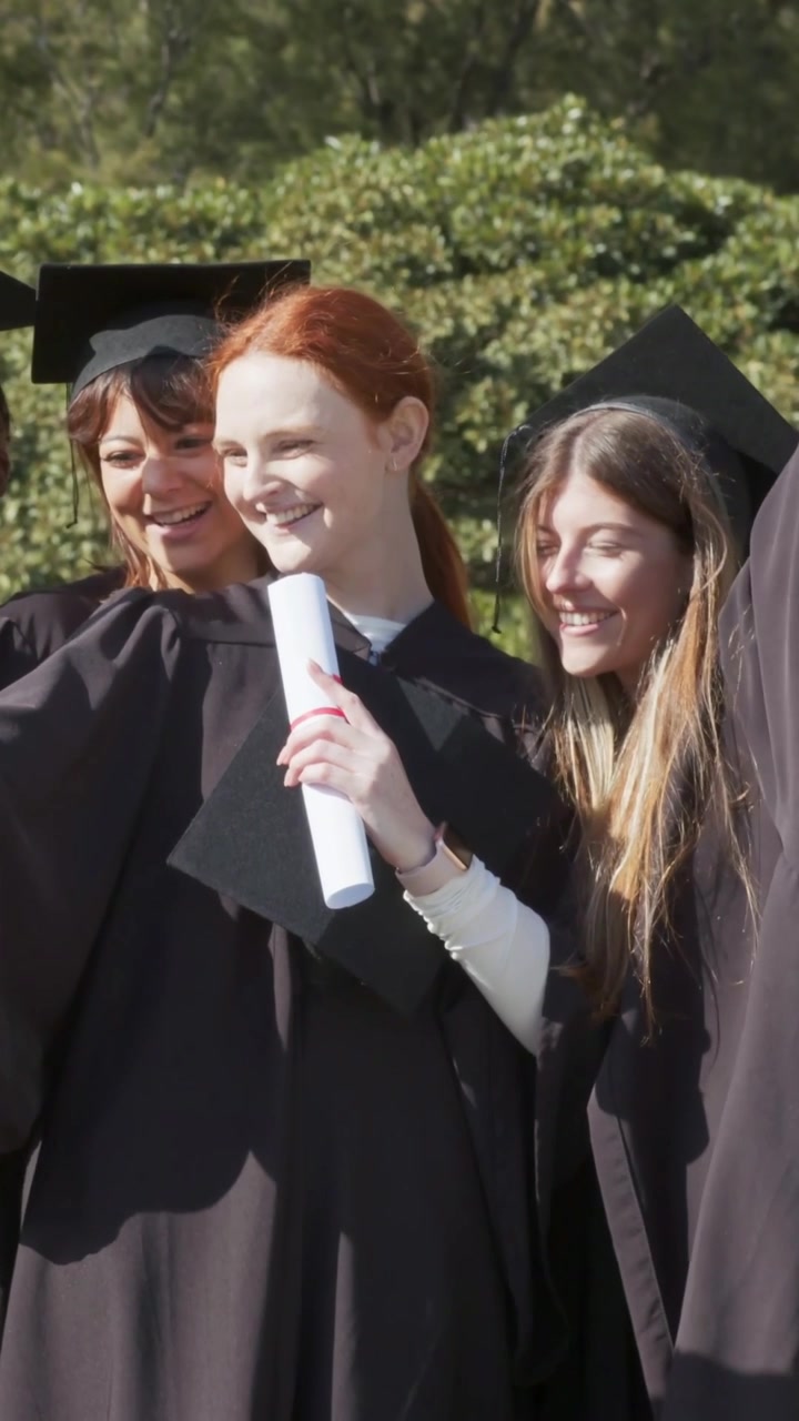 Video verticale: Tre laureati sorridenti che tengono i diplomi in toga nel campus, inclinati verso destra, festeggiando.
