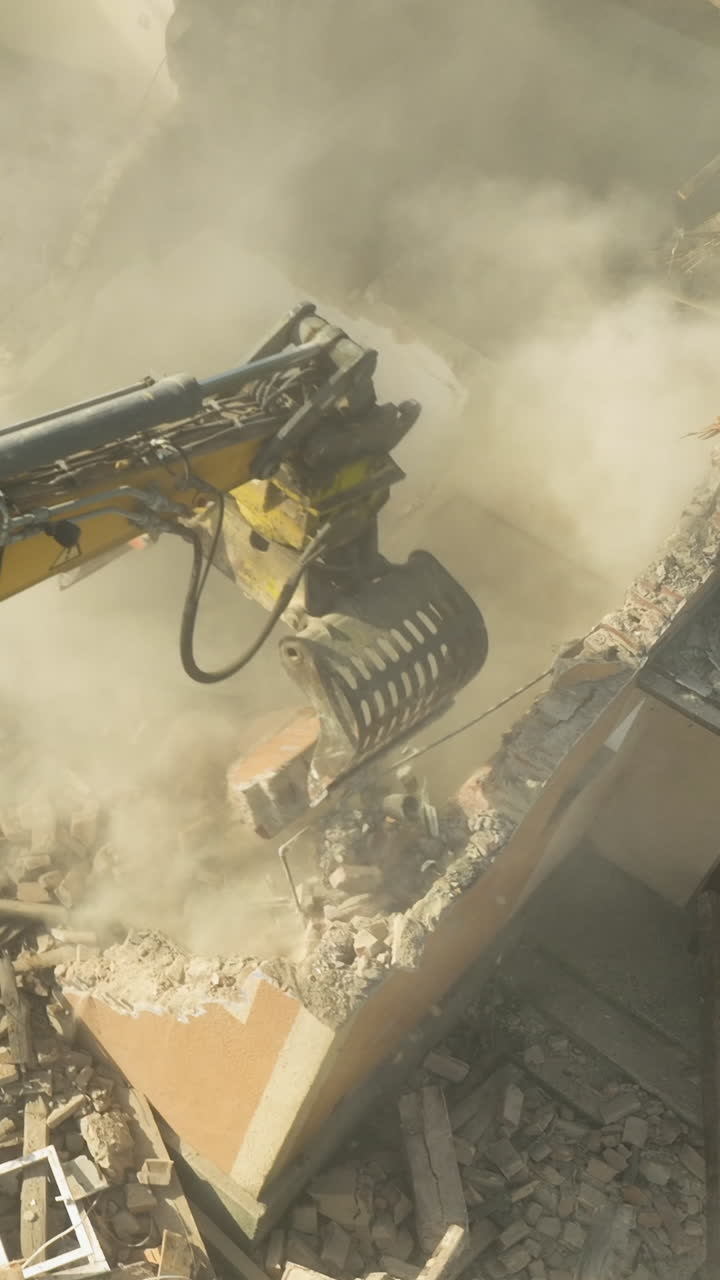 Une lourde pelle hydraulique écrase un mur de briques lors d'un processus de démolition, créant un énorme nuage de poussière. Gros plan cinématographique sur la rénovation urbaine et l'activité d'un chantier de construction.