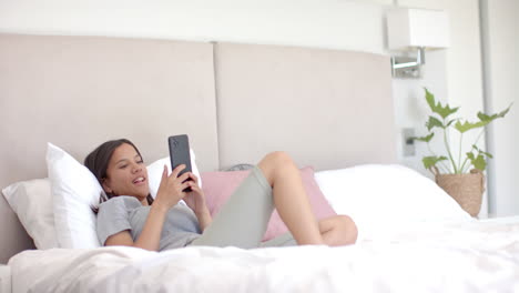 biracial teenage girl lying on bed at home, holding smartphone, with copy space